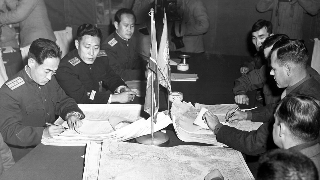 group of high-ranking officers at conference table signing documents with flags on table