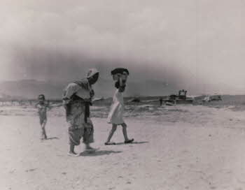 A old woman carrying a baby on her back, a young girl carrying laundry on her head, and a naked boy