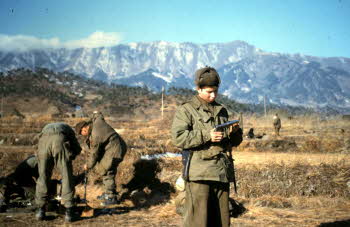 Pete Doyle looking at his 45 auto pistol after cleaning. Men in back cleaning weapons. Peak of hill 1243 to right