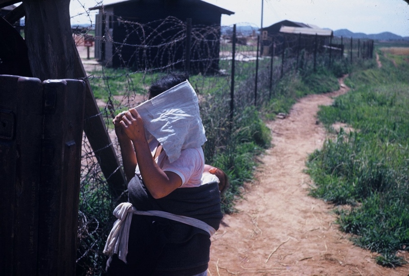 A mother carrying her baby while looking into the Army base from the other side of the barbed wire. Taken in Korea, 1953.